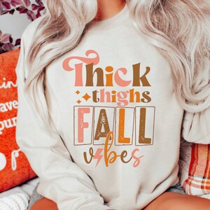 May include: A white sweatshirt with the text "Thick thighs fall vibes" in pink, brown, and orange lettering. The text is in a retro font style.