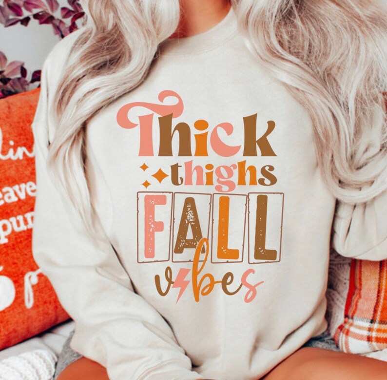 May include: A white sweatshirt with the text "Thick thighs fall vibes" in pink, brown, and orange lettering. The text is in a retro font style.
