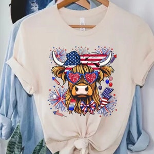 Puede incluir: Una camiseta blanca con un gráfico de una vaca de las Tierras Altas con gafas de sol en forma de corazón rojo, blanco y azul y una bandana con la bandera estadounidense. La vaca está rodeada de fuegos artificiales y otras imágenes patrióticas.