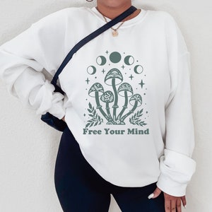 以下が含まれることがあります： 「Free Your Mind」というテキストと、キノコと月の緑色のグラフィックが描かれた白いスウェットシャツ。