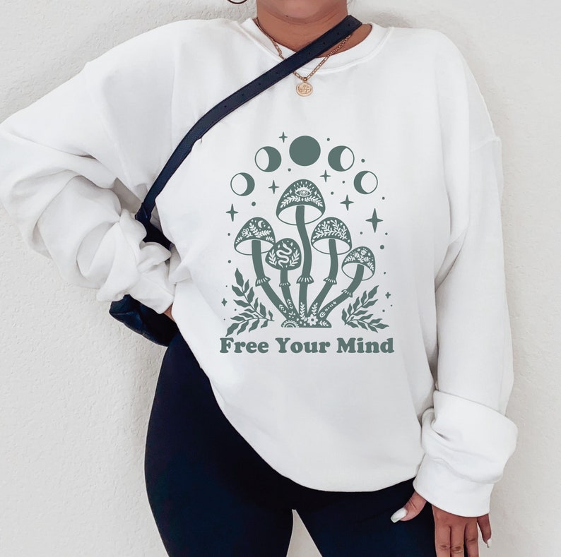 以下が含まれることがあります： 「Free Your Mind」というテキストと、キノコと月の緑色のグラフィックが描かれた白いスウェットシャツ。