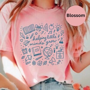 Puede incluir: Camiseta rosa claro con un gráfico azul que incluye un libro abierto, una manzana, una bombilla y el texto "helping little minds grow". La camiseta tiene cuello redondo y mangas cortas. La palabra "Blossom" está en un círculo rosa.