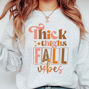 May include: A gray sweatshirt with the text "Thick thighs fall vibes" printed on the front in pink, brown, and orange. The text is in a retro font style.