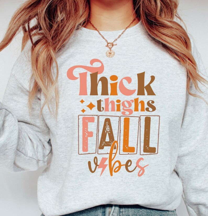 May include: A gray sweatshirt with the text "Thick thighs fall vibes" printed on the front in pink, brown, and orange. The text is in a retro font style.