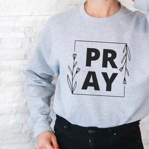 May include: A gray sweatshirt with the word "PRAY" in black lettering inside a square frame with floral accents.