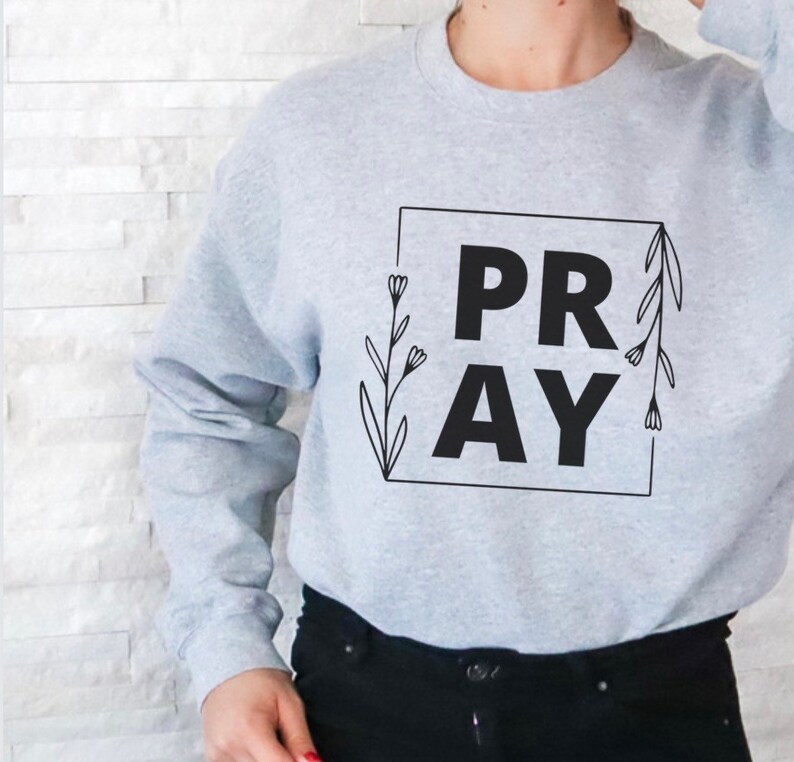 May include: A gray sweatshirt with the word "PRAY" in black lettering inside a square frame with floral accents.