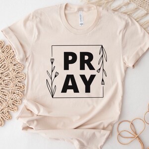 May include: A beige t-shirt with a black graphic that says "PRAY" with floral accents.