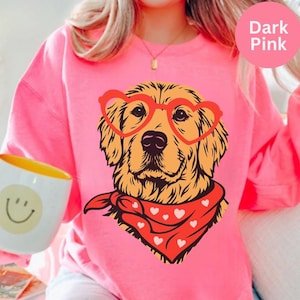 May include: A dark pink sweatshirt featuring a golden retriever wearing heart-shaped glasses and a red bandana with white hearts. The dog illustration is centered on the front. A white mug with a smiley face is visible on the left.