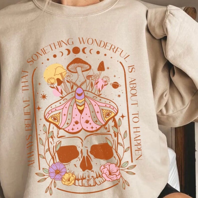 Op de afbeelding: Een beige sweatshirt met een grafische print van een schedel met een mot er bovenop. De grafiek is omringd door bloemen, paddenstoelen en sterren. De tekst "Geloof altijd dat er iets wonderlijks gaat gebeuren" is rond de grafiek gedrukt.