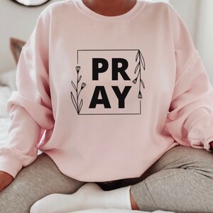 May include: A pink sweatshirt with the word "PRAY" in a black square with floral accents.
