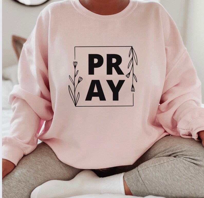 May include: A pink sweatshirt with the word "PRAY" in a black square with floral accents.