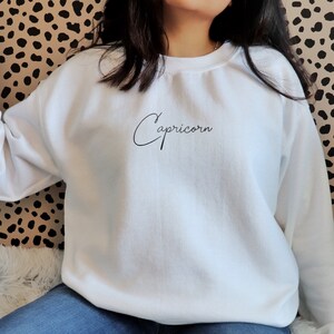 May include: White sweatshirt with the word "Capricorn" written in cursive on the front.