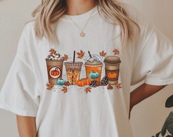 May include: White t-shirt with a graphic of four fall-themed coffee drinks with whipped cream, pumpkin spice, and fall leaves.