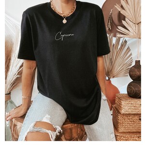May include: Black oversized t-shirt with the word "Capricorn" written in white cursive script.