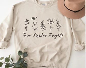 Può includere: Una felpa beige con un disegno floreale nero e il testo "Grow Positive Thoughts".