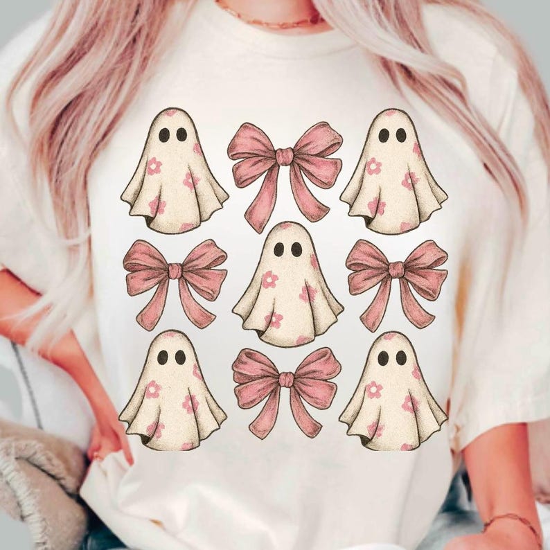 May include: Cream-colored t-shirt featuring a pattern of cartoon ghosts and pink bows. The ghosts have black eyes and are decorated with pink flowers. The bows are a solid pink color. The design is centered on the shirt.