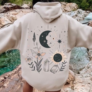 May include: A beige hoodie with a graphic print of a crescent moon, sun, crystals, and flowers. The print is in black, white, and gray.