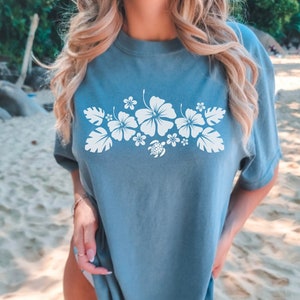 Peut inclure: Un t-shirt bleu clair avec un motif floral blanc représentant des fleurs d'hibiscus et une tortue. Le motif est centré sur la poitrine du t-shirt.