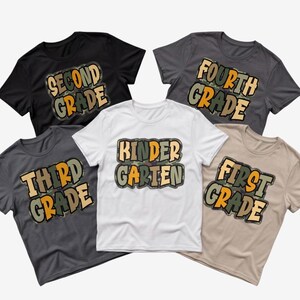 May include: A collection of five short-sleeved t-shirts in black, dark grey, white, and beige. Each shirt features a graphic print with camouflage-patterned lettering that reads Second Grade, Fourth Grade, Third Grade, Kindergarten, and First Grade.