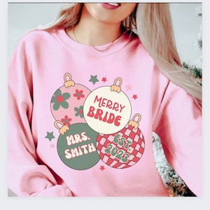 K&ouml;nnte beinhalten: Rosafarbener Pullover mit einem Weihnachtsbaumschmuck-Design. Die Baumschmuckst&uuml;cke haben den Text "Merry Bride", "Mrs. Smith" und "Est. 2023".