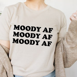 May include: A beige t-shirt with the text "MOODY AF" repeated three times in black.