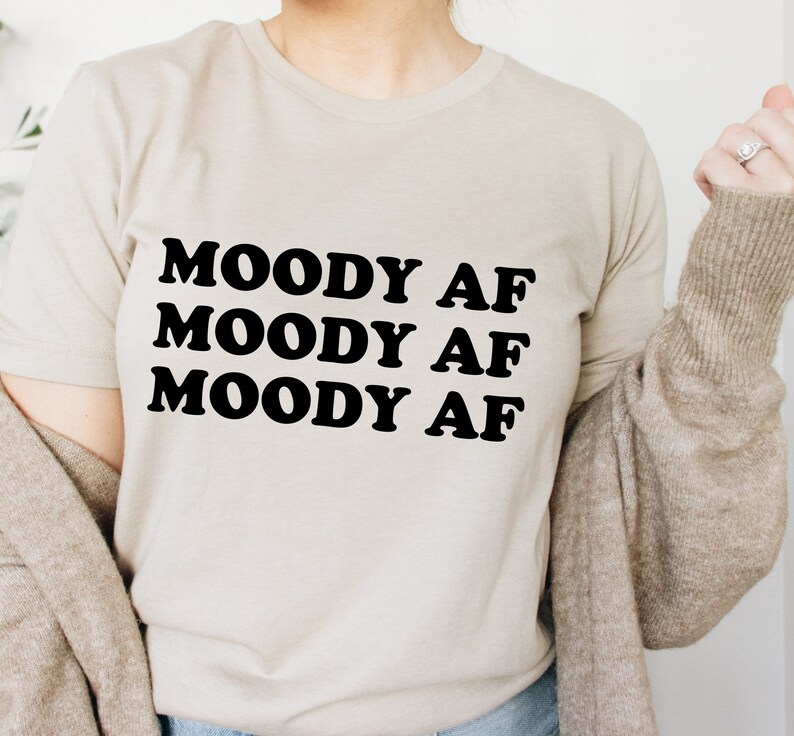 May include: A beige t-shirt with the text "MOODY AF" repeated three times in black.