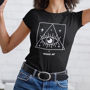 May include: Black t-shirt with a white graphic of an eye inside a triangle, surrounded by stars. The text "WOKE AF" is below the graphic. The shirt has a round neckline and short sleeves.