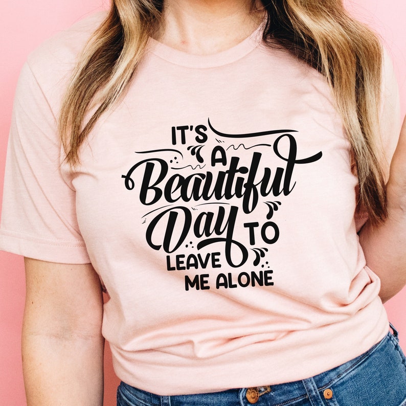 May include: A light pink t-shirt with the phrase "It's a Beautiful Day to Leave Me Alone" in black script. The shirt is made of a soft material and is suitable for casual wear.