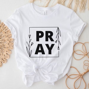 May include: White t-shirt with black text that says "PRAY" inside a square frame with floral accents.