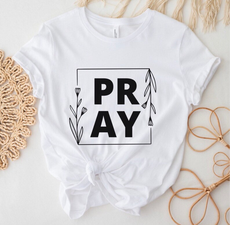 May include: White t-shirt with black text that says "PRAY" inside a square frame with floral accents.