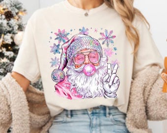 May include: A white t-shirt with a pink and white illustration of Santa Claus wearing sunglasses and blowing a bubble. The background is a watercolor design with snowflakes.