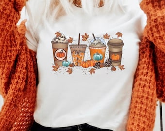 May include: White t-shirt with a graphic of four fall-themed coffee drinks with pumpkin spice, whipped cream, and fall leaves.