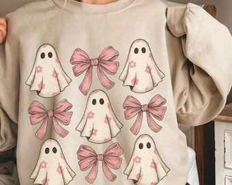 May include: Beige sweatshirt featuring a pattern of pink bows and ghost figures with pink flower details. The ghosts have black oval eyes. The design is centered on the front of the sweatshirt.