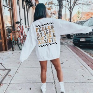 Peut inclure: Sweat-shirt blanc avec une bordure noire et blanche en damier autour du texte "Fueled by: Iced Coffee & Anxiety" en lettres brunes, beiges et blanches.