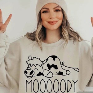 Peut inclure: Sweat-shirt beige avec un dessin de vache noir et blanc et le mot "MOOOOODY". Le sweat-shirt est de couleur neutre et a une coupe d&eacute;contract&eacute;e. Le dessin de la vache est un simple dessin au trait.