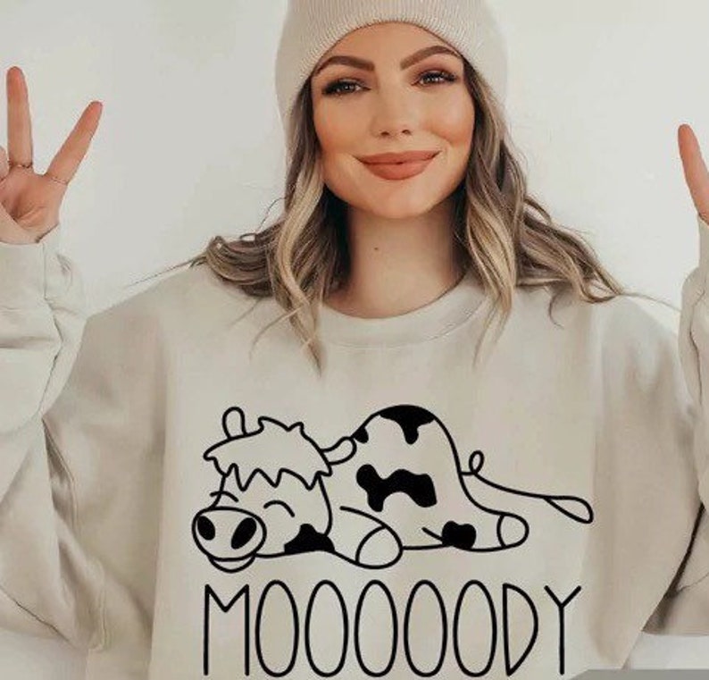 Peut inclure: Sweat-shirt beige avec un dessin de vache noir et blanc et le mot "MOOOOODY". Le sweat-shirt est de couleur neutre et a une coupe d&eacute;contract&eacute;e. Le dessin de la vache est un simple dessin au trait.