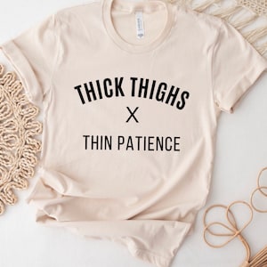 May include: A beige t-shirt with black text that reads "THICK THIGHS x THIN PATIENCE".