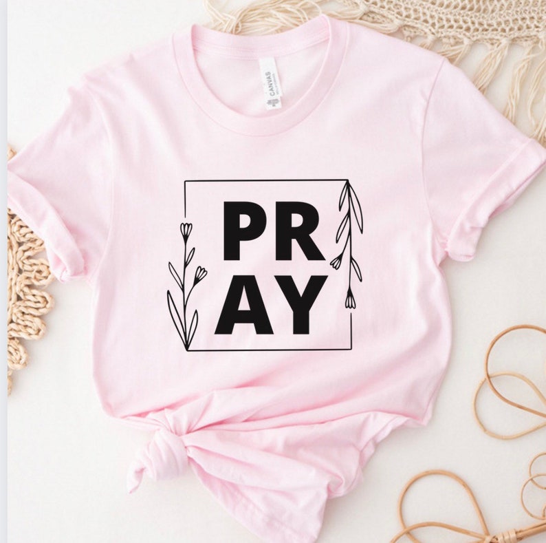 May include: A pink t-shirt with a black square graphic that says "PRAY" with floral accents.