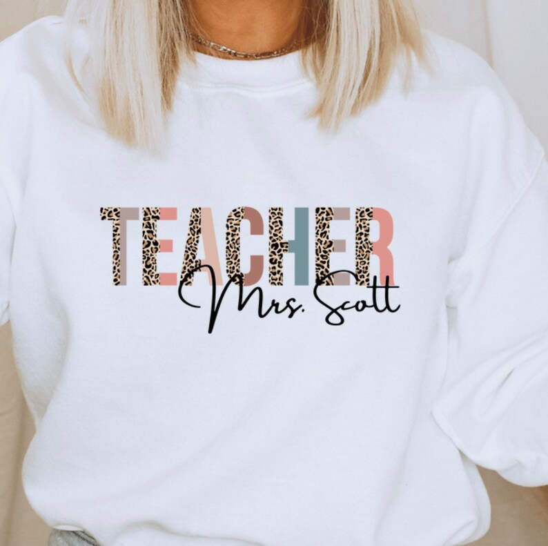 Pode incluir: Camisola branca com a palavra "TEACHER" em letras de estampa de leopardo com detalhes rosa, azul e marrom. O texto "Mrs. Scott" est&aacute; escrito em it&aacute;lico abaixo da palavra "TEACHER".