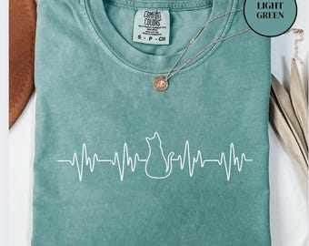 Può includere: T-shirt Comfort Colors verde chiaro con un design a silhouette di gatto bianco e linea del battito cardiaco. La maglietta ha girocollo e maniche corte. Visibile una collana d'argento con un ciondolo.