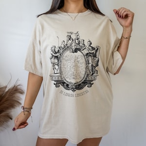 May include: An oversized beige t-shirt featuring a black and white graphic print of a vintage illustration with a crest and the text "In Libras Libertas".