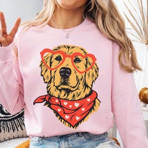 May include: A light pink sweatshirt featuring a golden retriever illustration. The dog wears heart-shaped glasses and a red bandana with white hearts. The design is centered on the front of the sweatshirt.