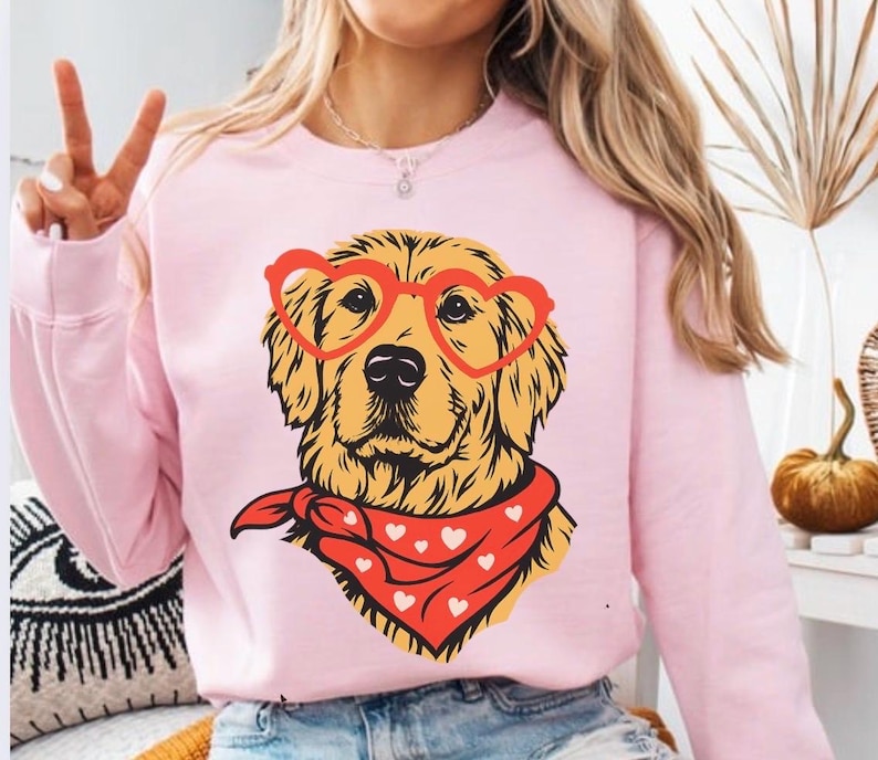 May include: A light pink sweatshirt featuring a golden retriever illustration. The dog wears heart-shaped glasses and a red bandana with white hearts. The design is centered on the front of the sweatshirt.