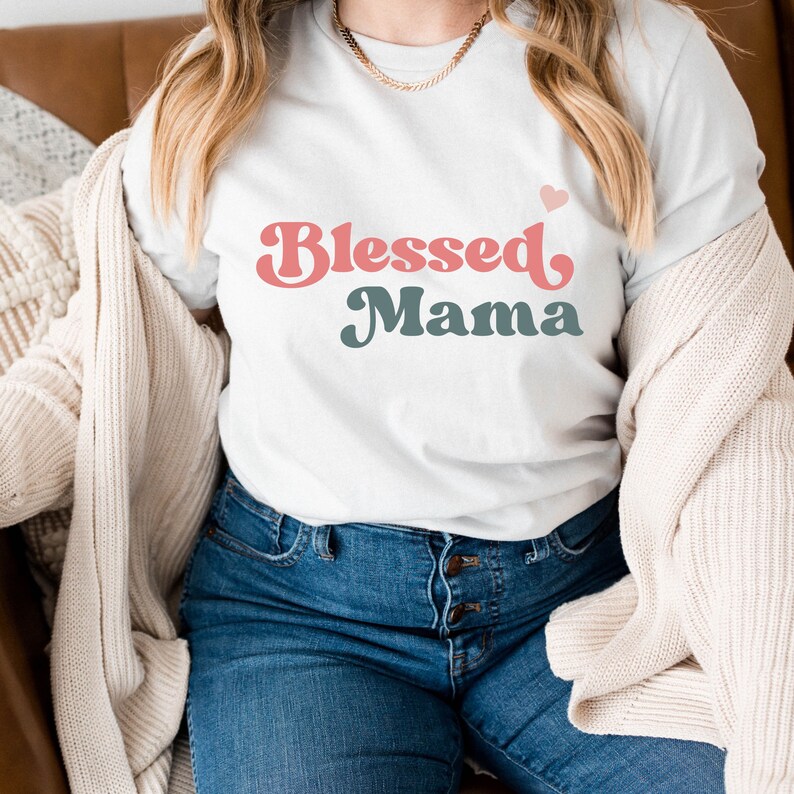 Puede incluir: Una camiseta blanca con el texto "Blessed Mama" en letras retro rosas y azules. Un peque&ntilde;o coraz&oacute;n rosa est&aacute; por encima del texto.