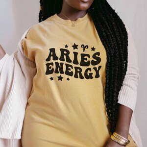 May include: Mustard yellow t-shirt with the words "ARIES ENERGY" in a retro font, with star and ram symbol accents. The shirt is a solid color and has a crew neck.
