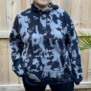 May include: A black hooded sweatshirt featuring a bleached, mottled gray and dark brown pattern throughout. The garment includes a front kangaroo pocket, a drawstring hood with black cords, and long sleeves with ribbed cuffs at the wrists.