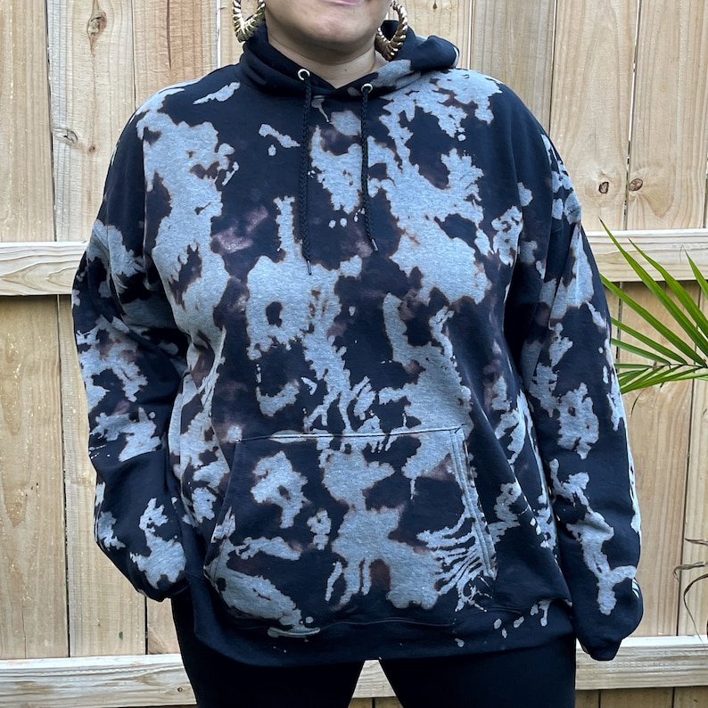 May include: A black hooded sweatshirt featuring a bleached, mottled gray and dark brown pattern throughout. The garment includes a front kangaroo pocket, a drawstring hood with black cords, and long sleeves with ribbed cuffs at the wrists.