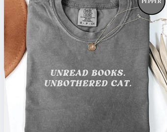 Może przedstawiać: Szary T-shirt z nadrukiem "Unread Books. Unbothered Cat." w kolorze białym. Koszulka ma postarzony wygląd i jest wykonana z miękkiej, wygodnej tkaniny.