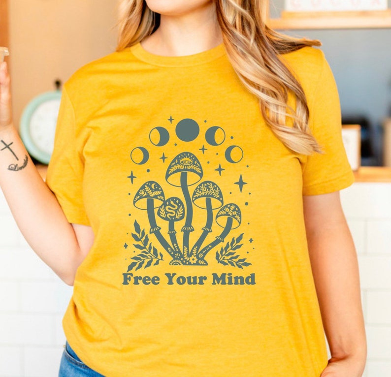 以下が含まれることがあります： 「Free Your Mind」というテキストと、キノコと三日月を描いたグラフィックがプリントされた黄色のTシャツです。
