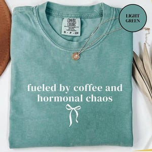 Puede incluir: Una camiseta verde claro Comfort Colors con el texto "fueled by coffee and hormonal chaos" en blanco. Un peque&ntilde;o gr&aacute;fico de lazo est&aacute; debajo del texto. Un collar plateado con un colgante est&aacute; en la camiseta.